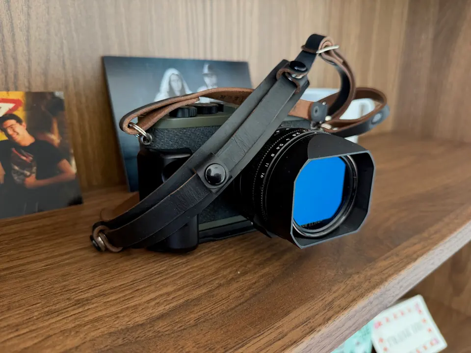 Leica Q2 Reporter Edition on a wooden shelf, its olive body fitted with a square vented hood holding a blue filter, a black leather strap draped across it, photographic prints propped behind