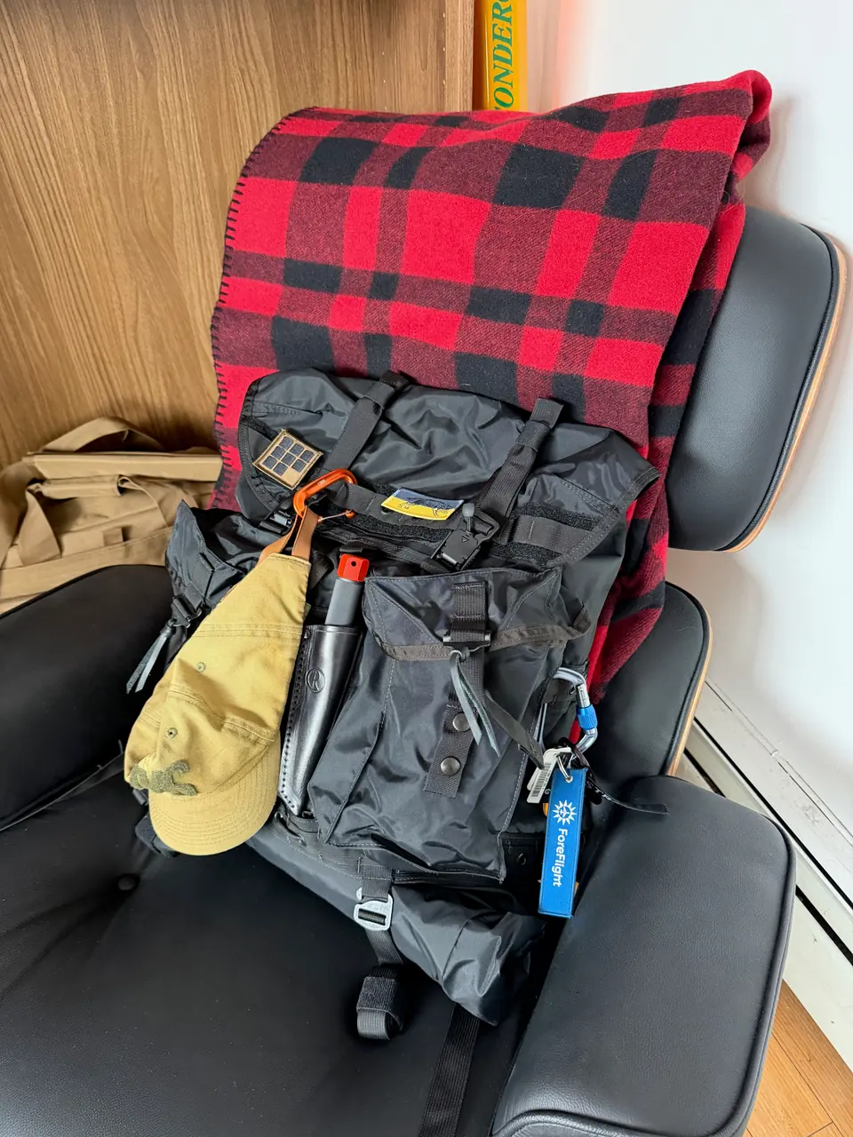 Black rucksack on a leather lounge chair, a red and black buffalo check wool blanket draped behind it, a tan cap and a carabiner with a ForeFlight tag clipped to the front